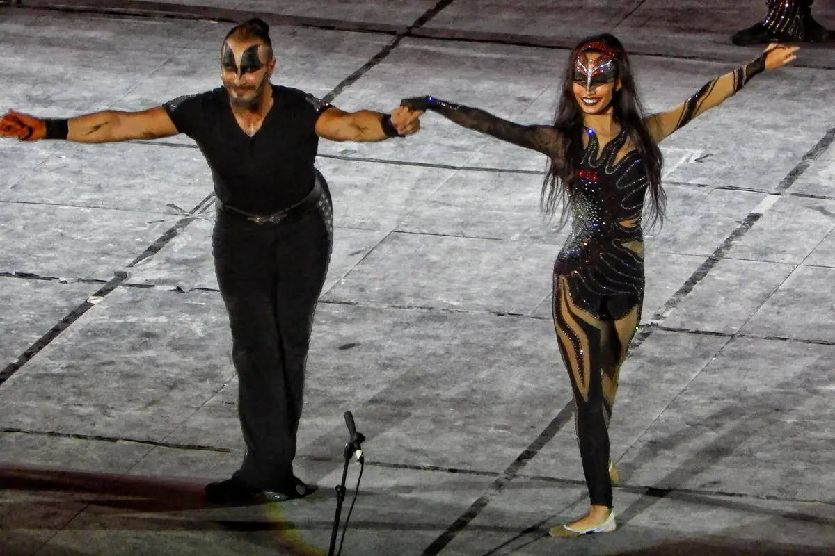 Two Fire of Anatolia lead dancers in black stage costumes and painted masks, holding hands and taking a bow